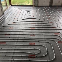 DSI INSTALLATIONS SPÓŁKA Z OGRANICZONĄ ODPOWIEDZIALNOŚCIĄ - Białe rury ogrzewania podłogowego ułożone spiralnie na szarej macie izolacyjnej w surowym wnętrzu z oknem. Widoczne mocowania rur i logo producenta na macie.