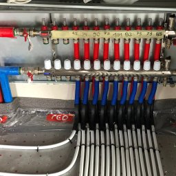 DSI INSTALLATIONS SPÓŁKA Z OGRANICZONĄ ODPOWIEDZIALNOŚCIĄ - Rozdzielacz ogrzewania podłogowego z rurami w kolorach czerwonym i niebieskim, zamontowany w metalowej szafce, z widocznymi numerami obwodów na taśmie.