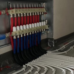 DSI INSTALLATIONS SPÓŁKA Z OGRANICZONĄ ODPOWIEDZIALNOŚCIĄ - Rozdzielacz ogrzewania podłogowego z rurami PEX w kolorach: czerwonym, niebieskim i czarnym, zamontowany na ścianie, z widocznymi numerami na taśmie.
