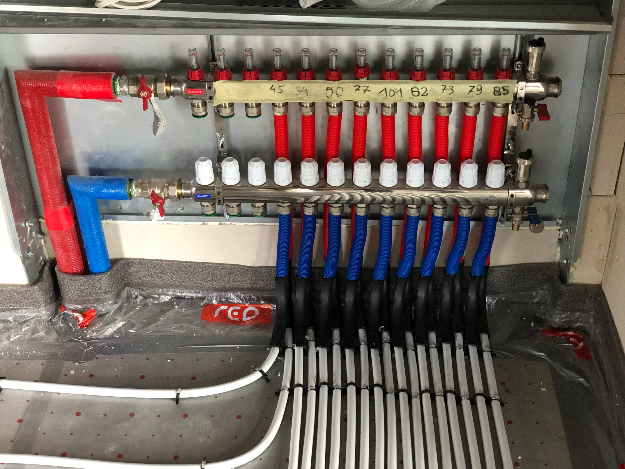 Rozdzielacz ogrzewania podłogowego z rurami w kolorach czerwonym i niebieskim, zamontowany w metalowej szafce, z widocznymi numerami obwodów na taśmie.
