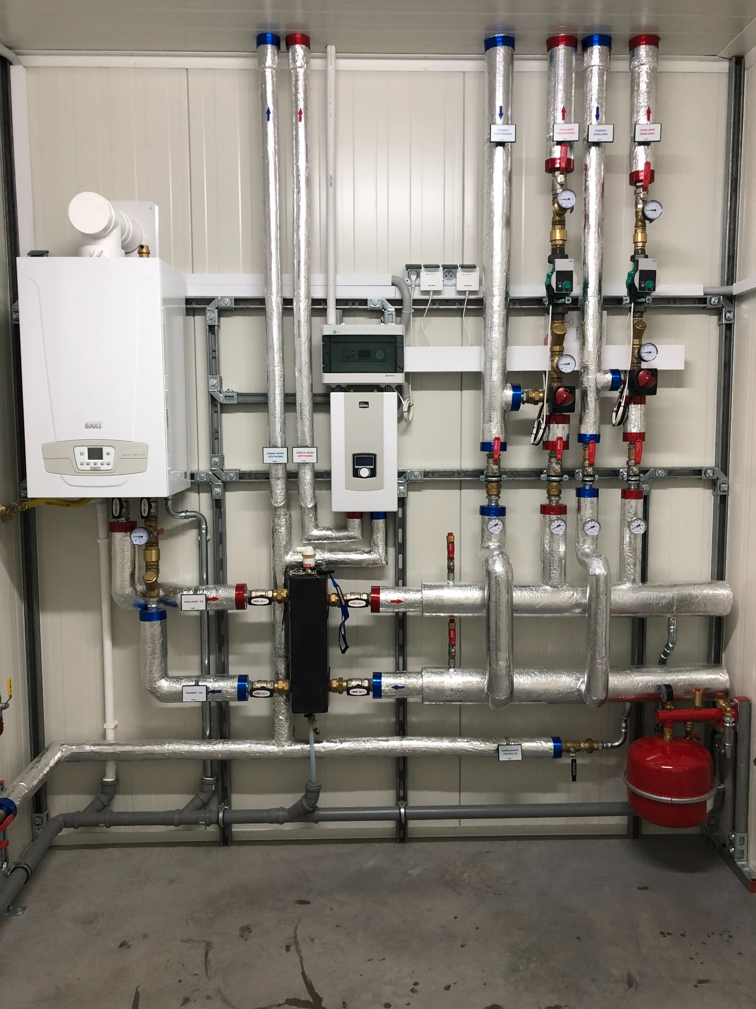 Kompletna instalacja grzewcza z kotłem gazowym, rurami w izolacji, zaworami i manometrami, zamontowana na ścianie w pomieszczeniu technicznym. Widoczne elementy kotłowni.