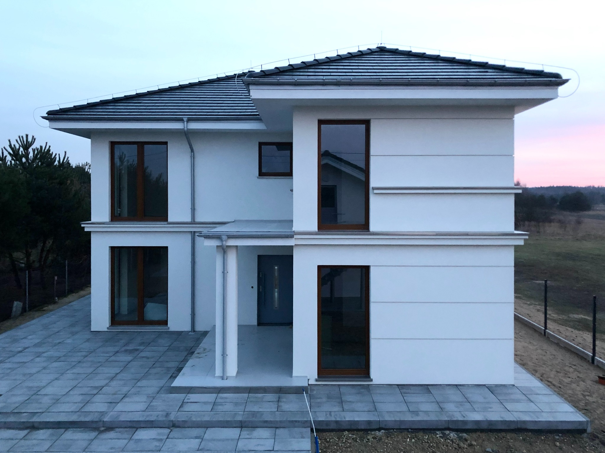 Dwukondygnacyjny, biały dom z ciemną dachówką i brązowymi ramami okiennymi, widziany z zewnątrz, z kostką brukową przed wejściem i fragmentem ogrodzenia w tle.