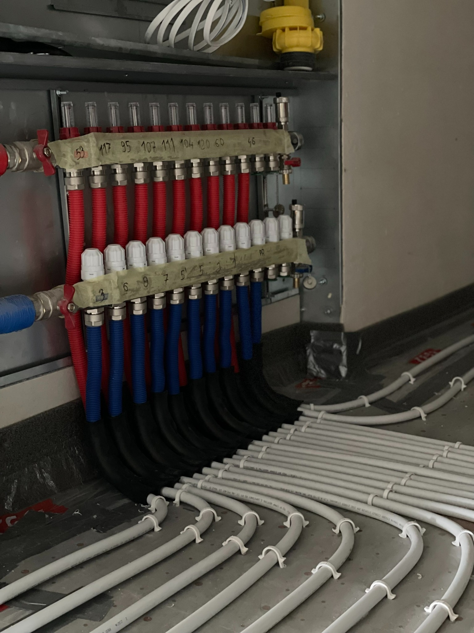 Rozdzielacz ogrzewania podłogowego z rurami PEX w kolorach: czerwonym, niebieskim i czarnym, zamontowany na ścianie, z widocznymi numerami na taśmie.