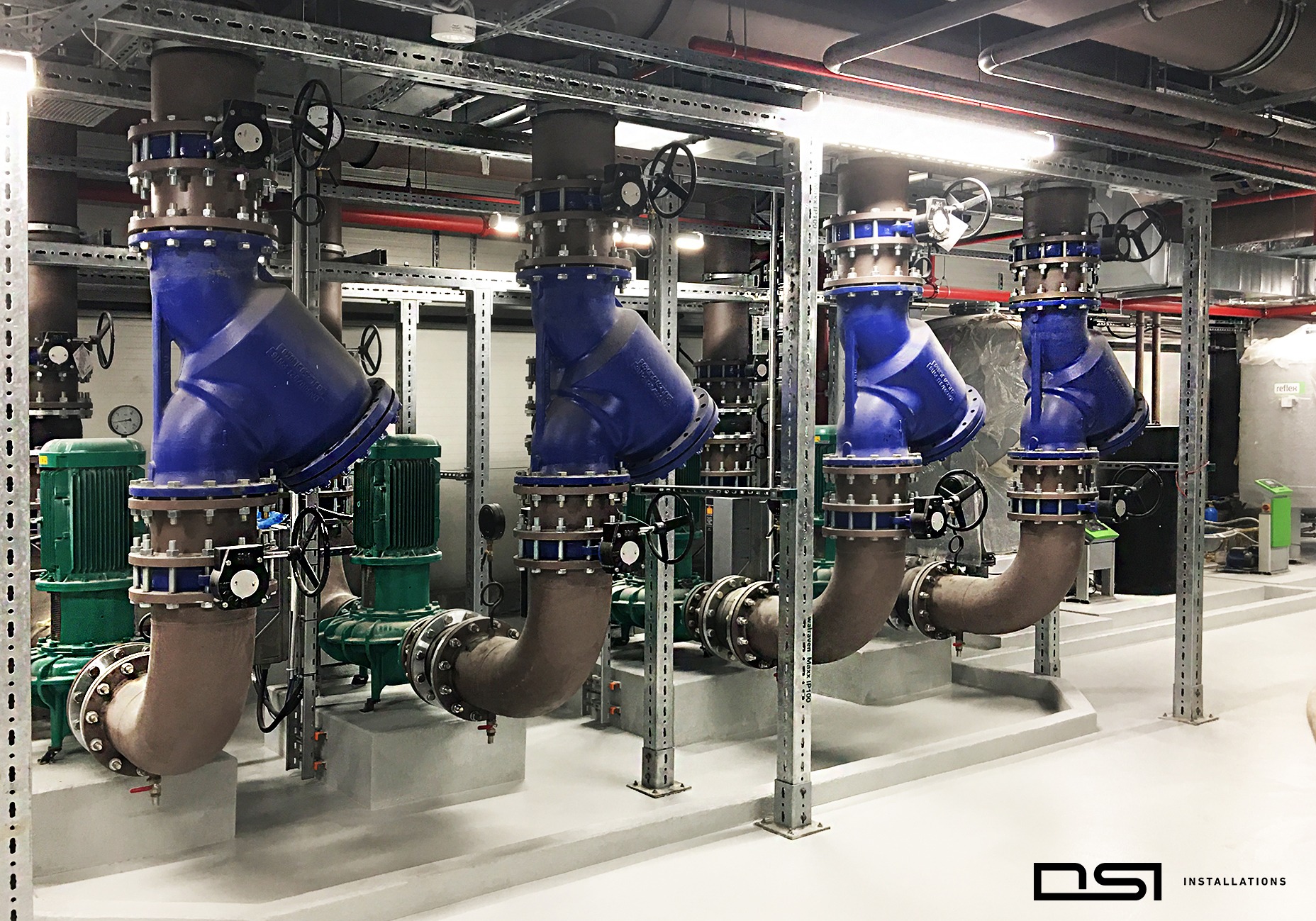 Rząd niebieskich zaworów i brązowych rur w instalacji przemysłowej, z zielonymi silnikami i metalową konstrukcją wsporczą. Oświetlenie sufitowe podkreśla czystość i porządek.
