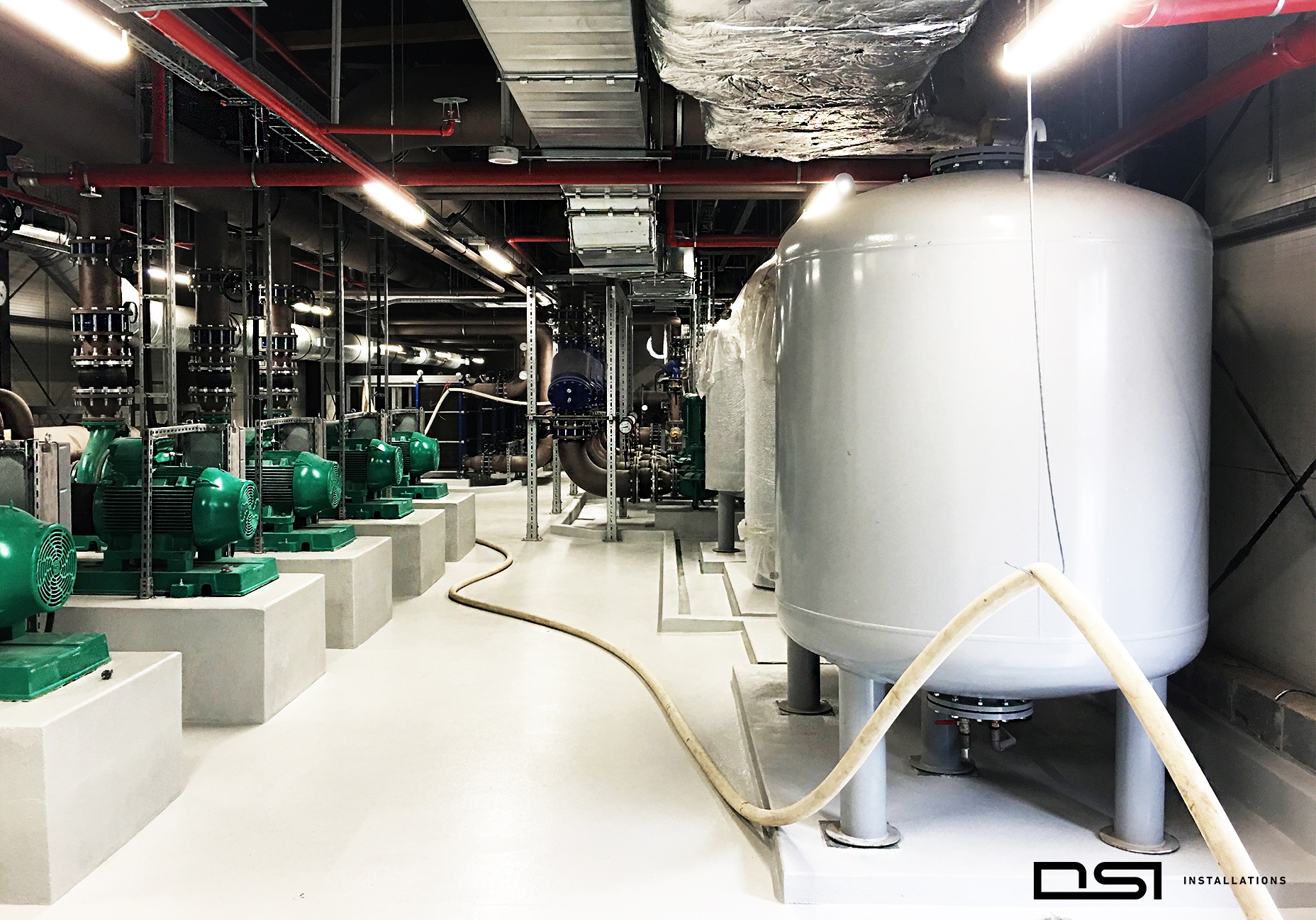 Nowoczesna kotłownia z szeregiem zielonych pomp i białych zbiorników ciśnieniowych, widoczne rury i instalacje pod sufitem. Jasne oświetlenie, czysta posadzka.