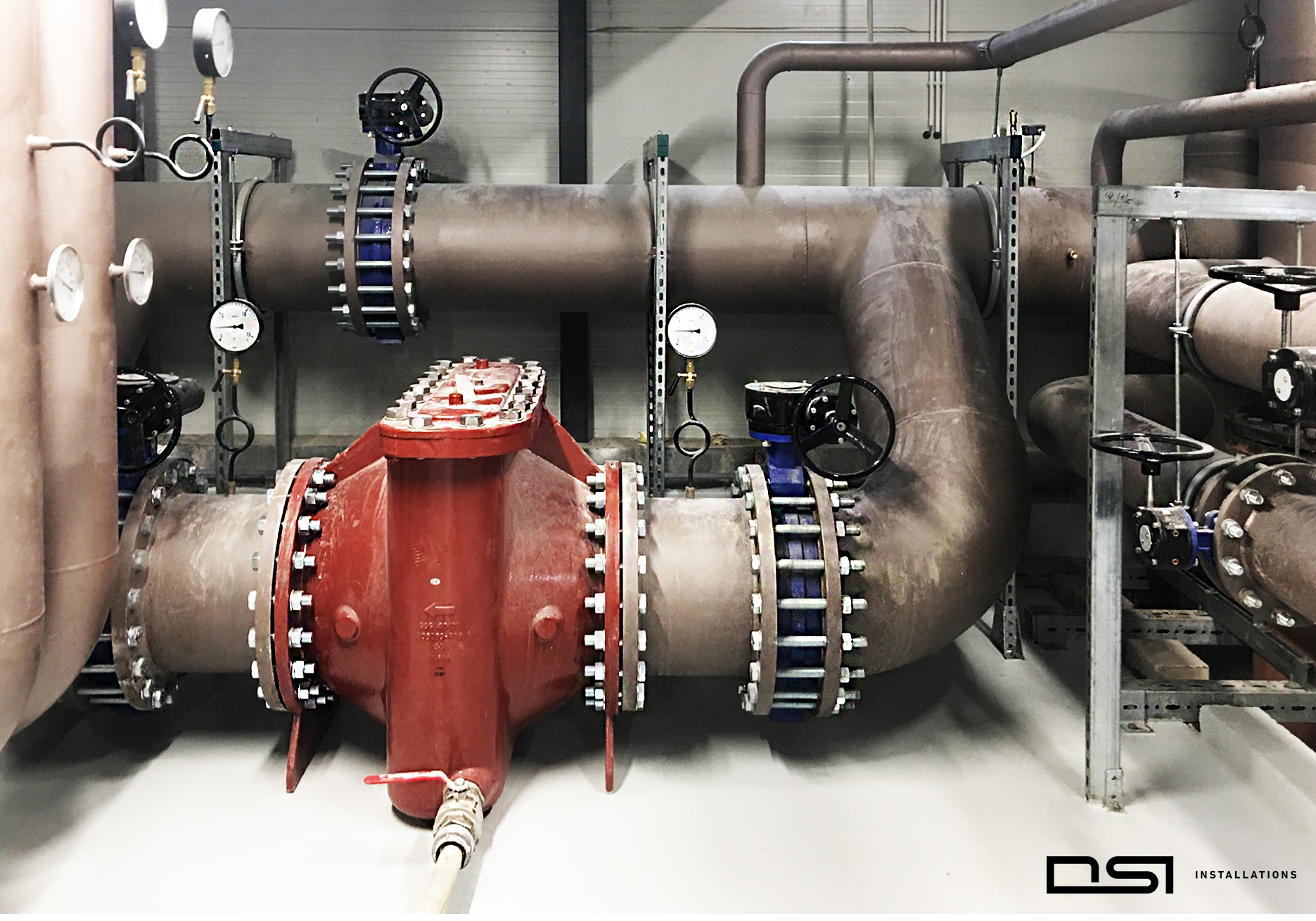 Złożona instalacja przemysłowa z rur, zaworów i manometrów. Czerwony zawór centralny z licznymi śrubami. Widoczne elementy mocujące i fragmenty konstrukcji wsporczej.