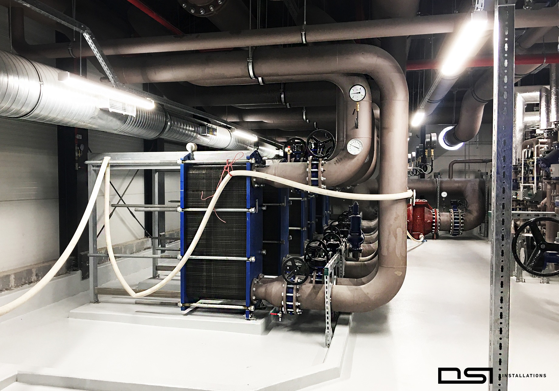 Węzeł cieplny z wymiennikiem płytowym, rurami, zaworami i manometrami w kotłowni. Widoczne elementy instalacji w przemysłowym wnętrzu.