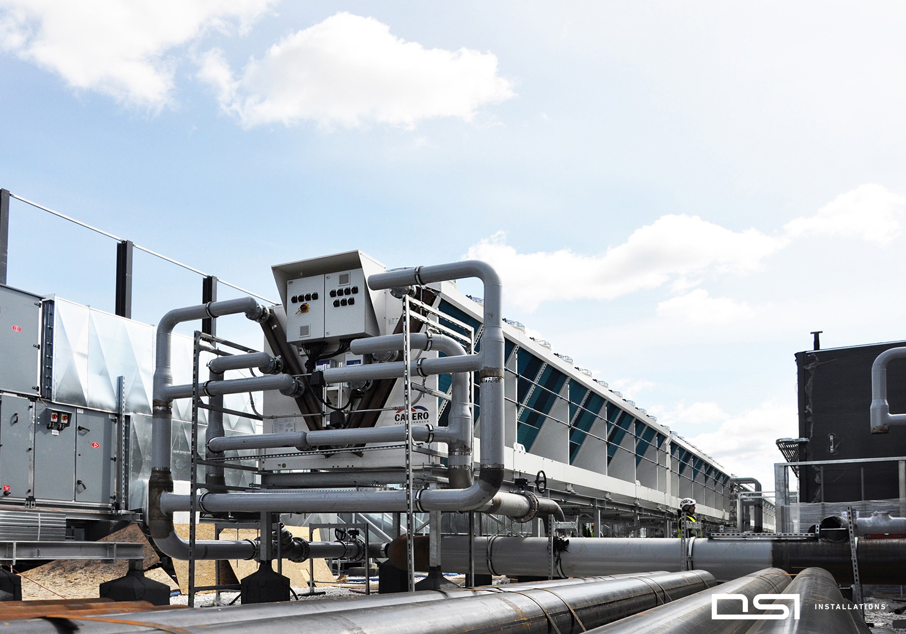 Instalacja przemysłowa chłodnicza na dachu budynku, widoczne rury, wentylatory i elementy sterujące. Błękitne niebo z chmurami w tle. Widok z dołu.