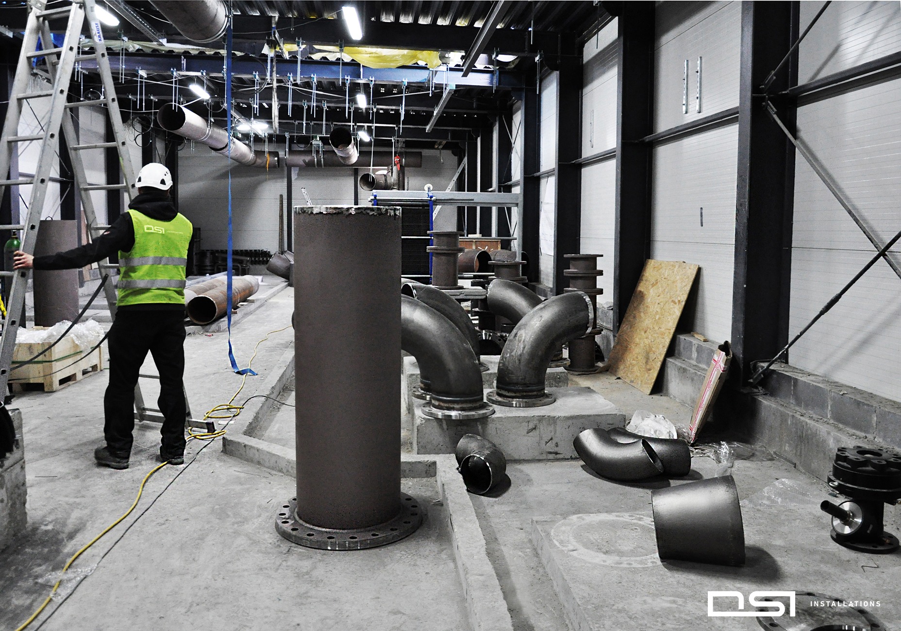 Montaż rur przemysłowych w hali: pracownik w kasku i kamizelce odblaskowej przy drabinie, liczne rury z kołnierzami na podłodze, surowy stan wykończenia.