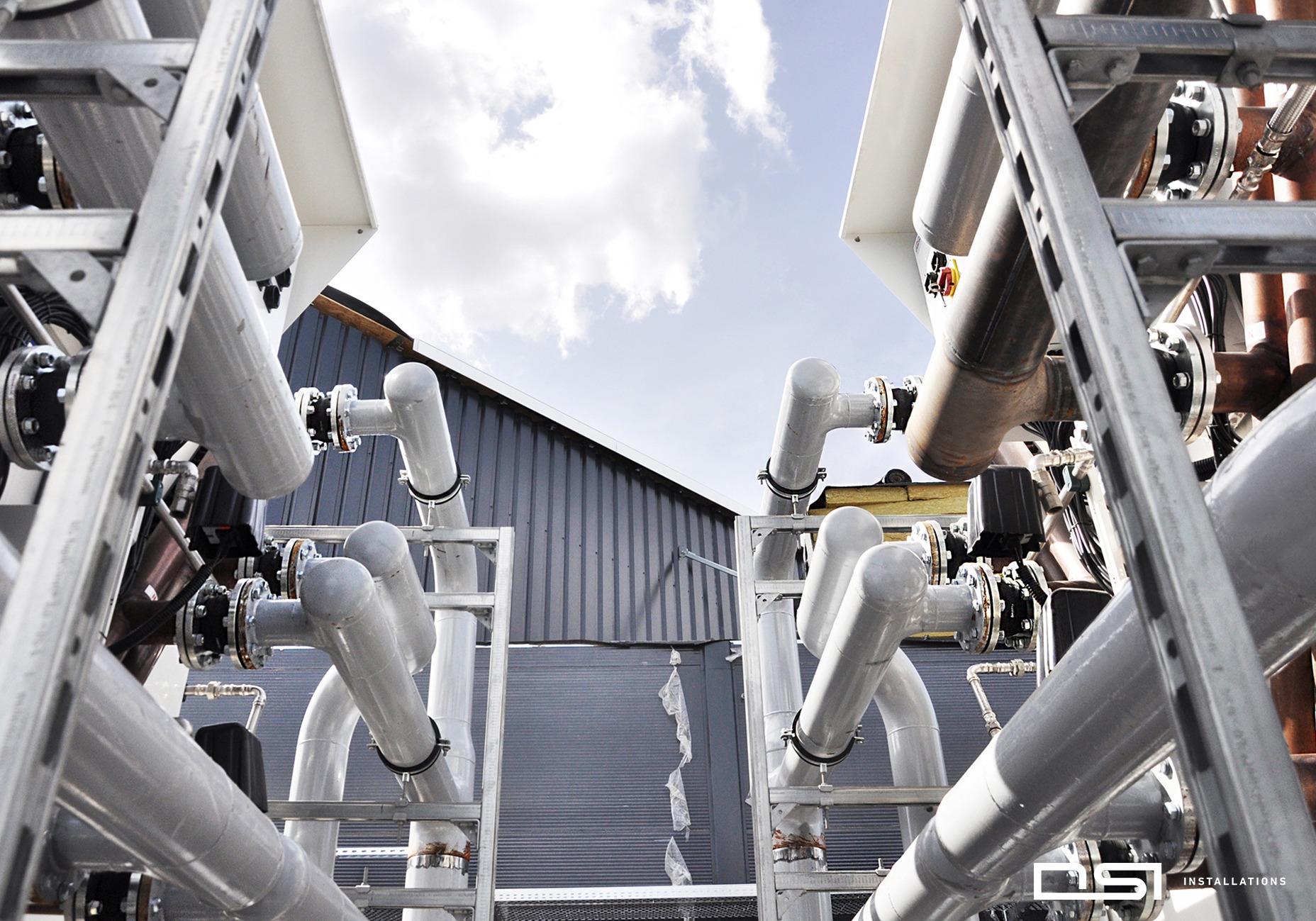 Kompleksowa instalacja przemysłowa z rurami w kolorze szarym, z widocznymi elementami mocującymi i fragmentem nieba w tle. Ujęcie z dołu, podkreślające złożoność konstrukcji.