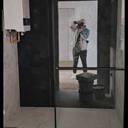 Vladyslav Panchenko RemExpert - Nowoczesna łazienka z czarną ścianą i kabiną prysznicową typu walk-in. W lustrze odbija się osoba robiąca zdjęcie. Widoczny bojler i elementy instalacji.