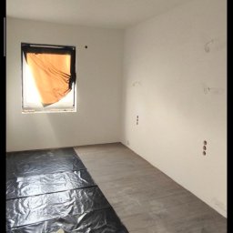 Vladyslav Panchenko RemExpert - Pokój w trakcie malowania: podłoga zabezpieczona folią, wałek i kuweta na folii, ściany przygotowane do malowania, okno zasłonięte folią. Widoczne gniazdka elektryczne.