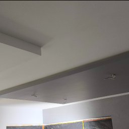 Dwupoziomowy sufit podwieszany z otworami na kable oświetleniowe, pomalowany na biało i szaro, w jasnym wnętrzu. Widoczne fragmenty ścian i okna zabezpieczonego folią.