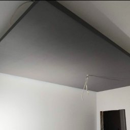 Szary sufit podwieszany z widocznym kablem, zamontowany w rogu białego pomieszczenia. Minimalistyczne wykończenie wnętrza, widok z dołu.
