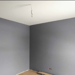 Świeżo pomalowane na szaro wnętrze pokoju z białym sufitem i widocznym oknem. Na podłodze leżą panele. Widoczny kabel zwisający z sufitu.