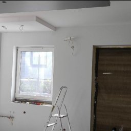 Vladyslav Panchenko RemExpert - Wnętrze w trakcie prac wykończeniowych: białe ściany, sufit podwieszany z szarą wnęką, okno z drabinką, widoczne przewody elektryczne i ślady po gładzi.