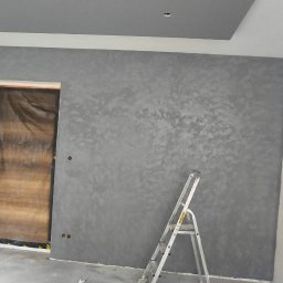 REMEXPERT - Ściana po dekoracyjnym malowaniu w odcieniach szarości, z widoczną fakturą. W tle zabezpieczone drzwi i drabina malarska. Sufit z podwieszanym elementem.