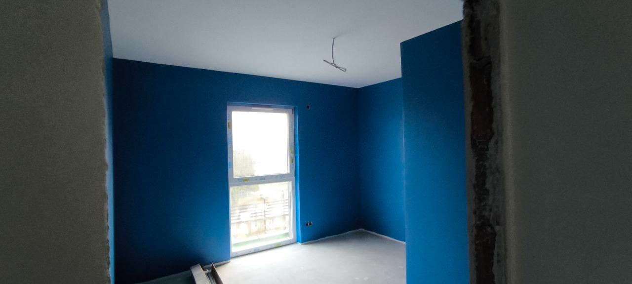 Pomalowane na niebiesko wnętrze pokoju z oknem. Widoczne ślady po remoncie na framugach. Na suficie wystają kable. Podłoga szara, surowa. Ściany w intensywnym odcieniu niebieskiego.
