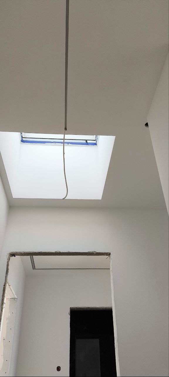 Białe wnętrze w trakcie prac wykończeniowych: sufit z oknem dachowym zabezpieczonym taśmą, widoczne przewody, przygotowane otwory drzwiowe.