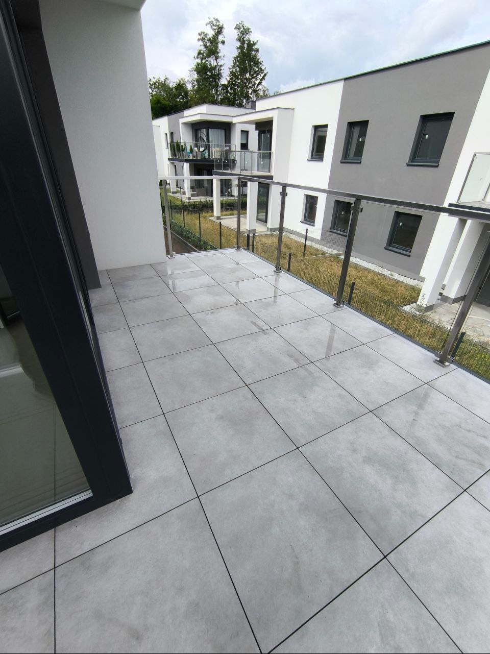 Balkon z szarymi płytkami, nowoczesne balustrady ze stali nierdzewnej i szkła, widok na budynek w tle. Minimalistyczny design, jasne otoczenie.