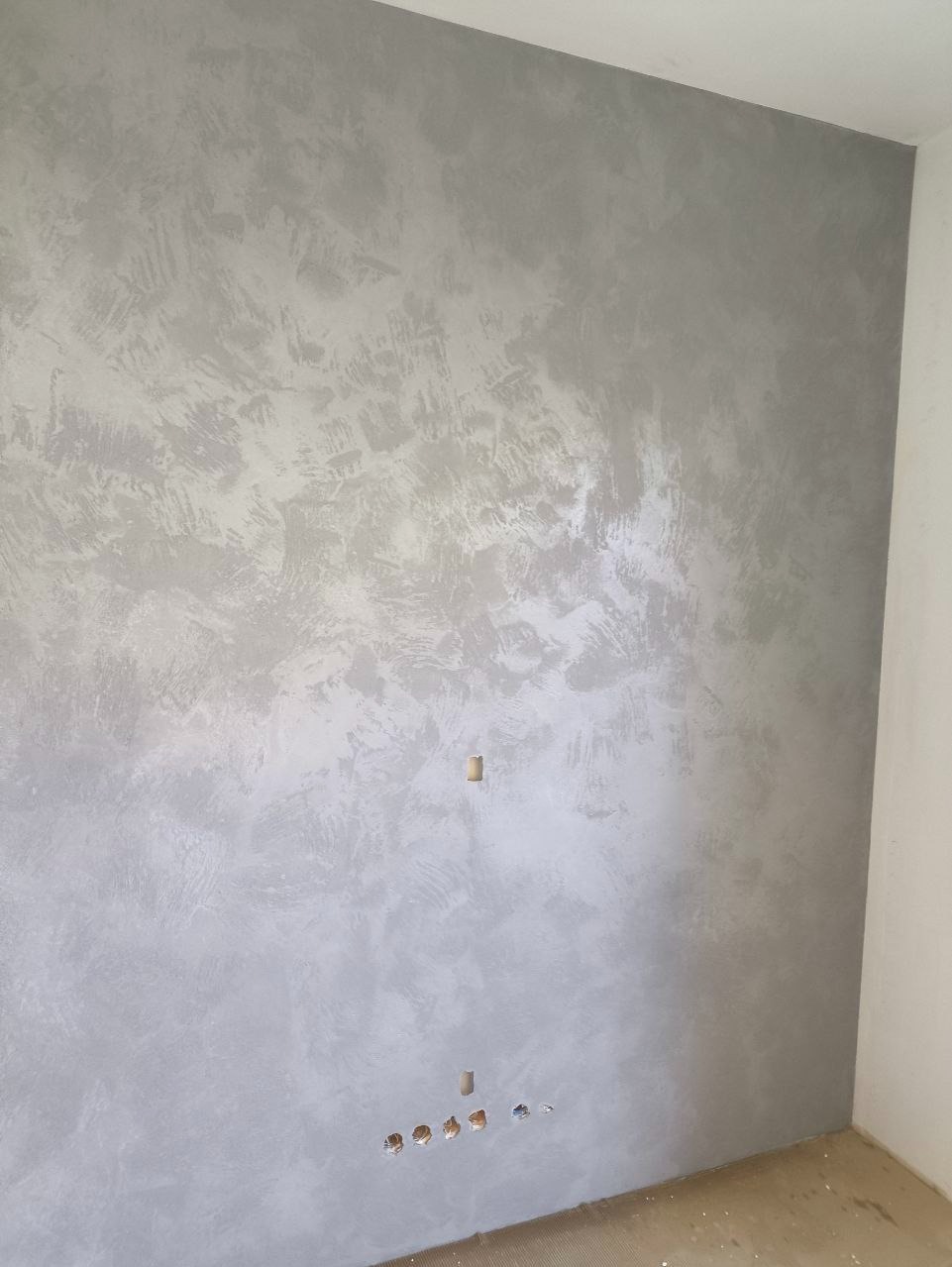 Ściana z dekoracyjnym, srebrzystoszarym tynkiem strukturalnym, widoczne gniazdka elektryczne i otwór na kabel. Minimalistyczny design, surowe wykończenie.