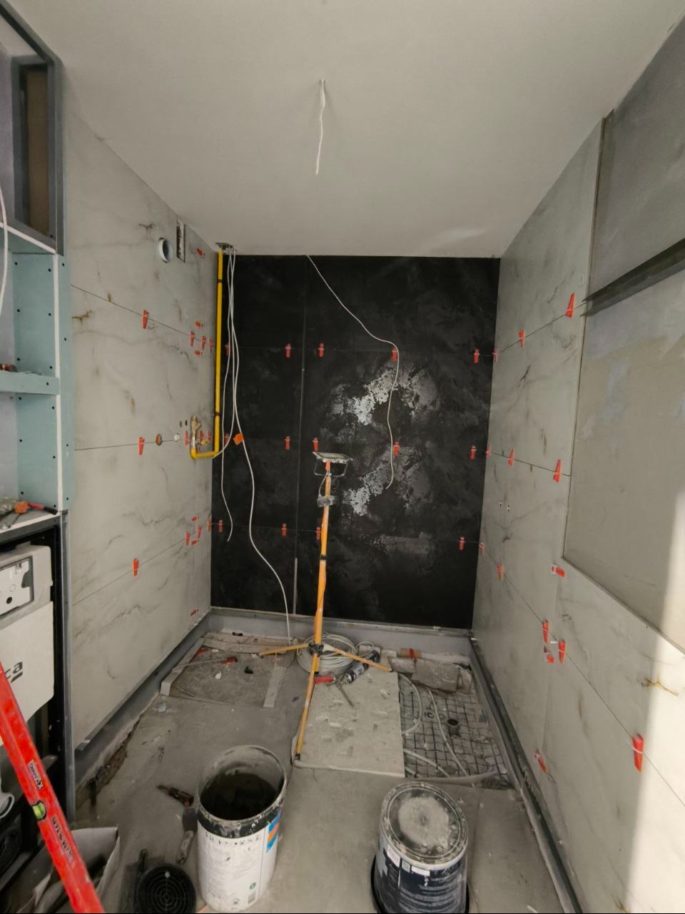 Łazienka w trakcie prac: biało-czarne płytki z krzyżykami dystansowymi, widoczne rury, lampa budowlana i wiaderka z zaprawą. Etap kładzenia płytek.