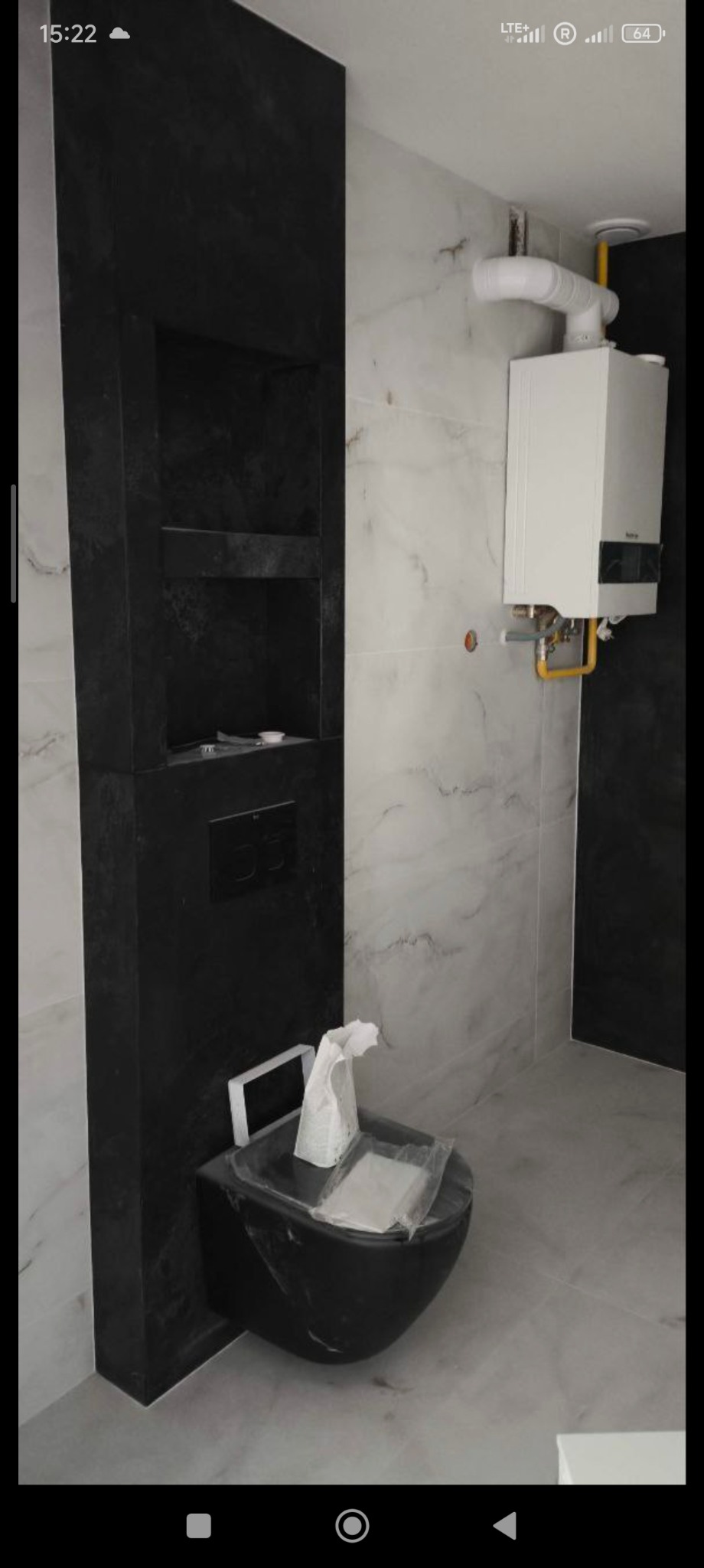 Wykończona łazienka z czarną armaturą: wisząca toaleta i wnęka z półkami. Na ścianach białe płytki imitujące marmur, widoczny bojler. Minimalistyczny design.