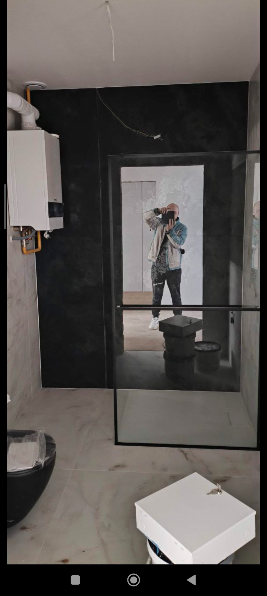 Nowoczesna łazienka z czarną ścianą i kabiną prysznicową typu walk-in. W lustrze odbija się osoba robiąca zdjęcie. Widoczny bojler i elementy instalacji.