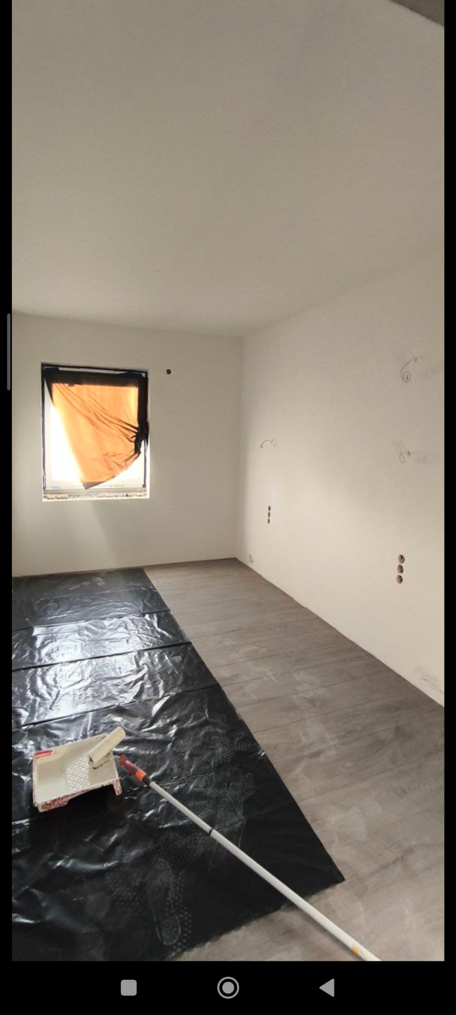 Pokój w trakcie malowania: podłoga zabezpieczona folią, wałek i kuweta na folii, ściany przygotowane do malowania, okno zasłonięte folią. Widoczne gniazdka elektryczne.
