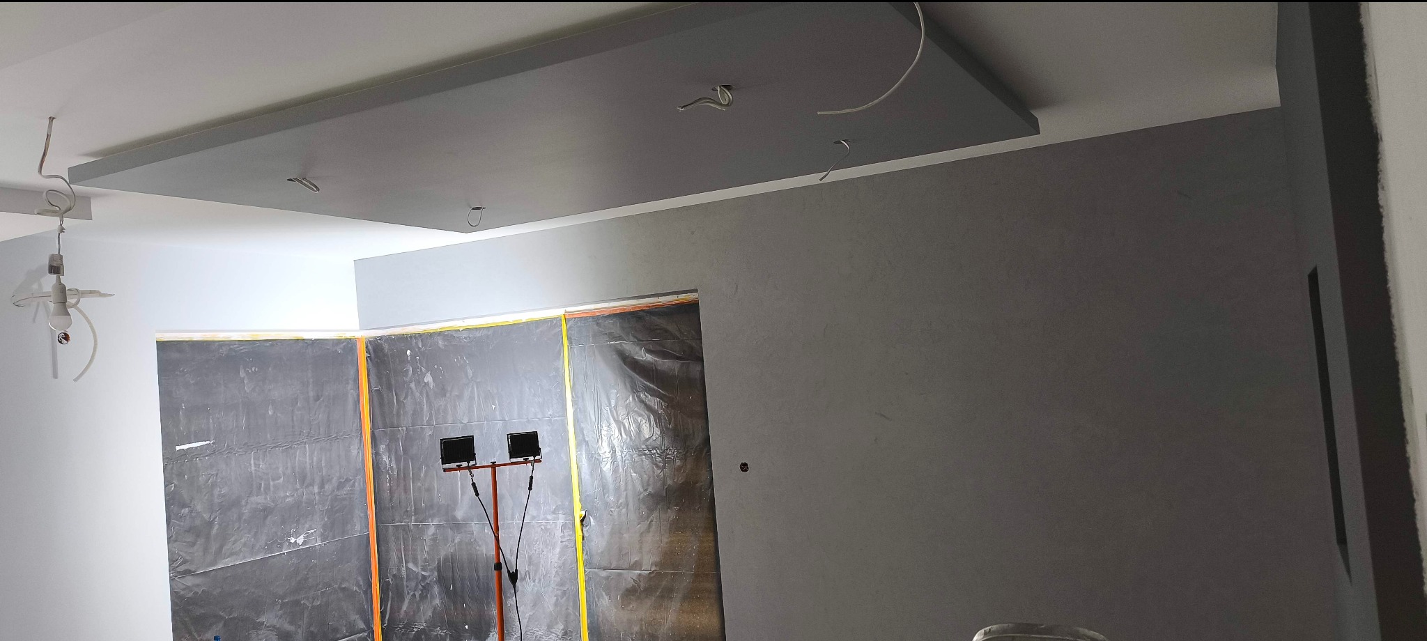 Szara ściana z zamontowaną płytą karton-gips na suficie. Zabezpieczone folią drzwi i lampa budowlana. W tle biała ściana z wiszącą żarówką.