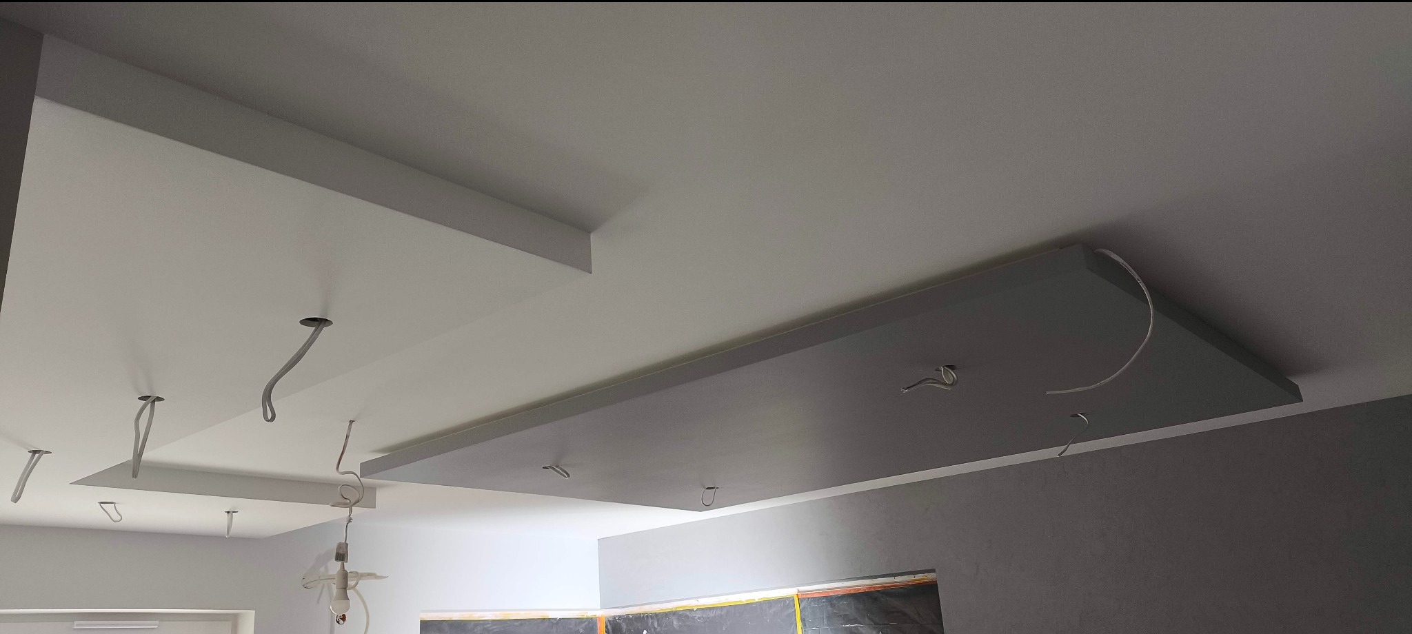 Dwupoziomowy sufit podwieszany z otworami na kable oświetleniowe, pomalowany na biało i szaro, w jasnym wnętrzu. Widoczne fragmenty ścian i okna zabezpieczonego folią.