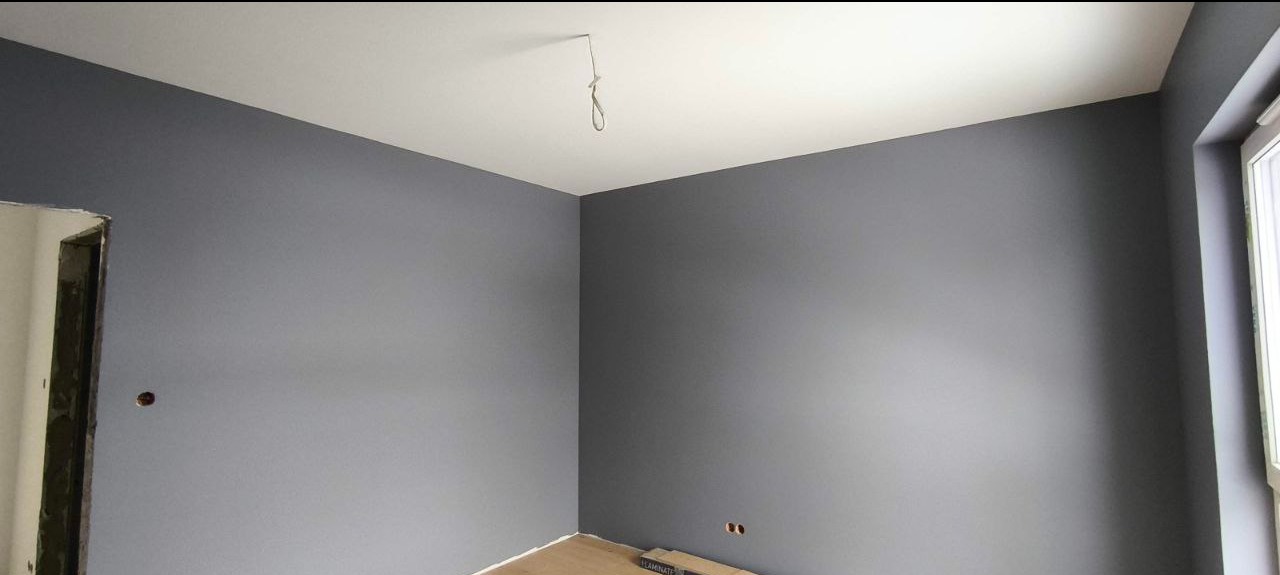 Świeżo pomalowane na szaro wnętrze pokoju z białym sufitem i widocznym oknem. Na podłodze leżą panele. Widoczny kabel zwisający z sufitu.