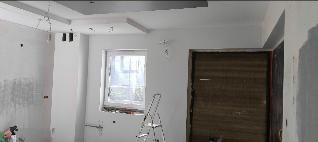 Wnętrze w trakcie prac wykończeniowych: białe ściany, sufit podwieszany z szarą wnęką, okno z drabinką, widoczne przewody elektryczne i ślady po gładzi.