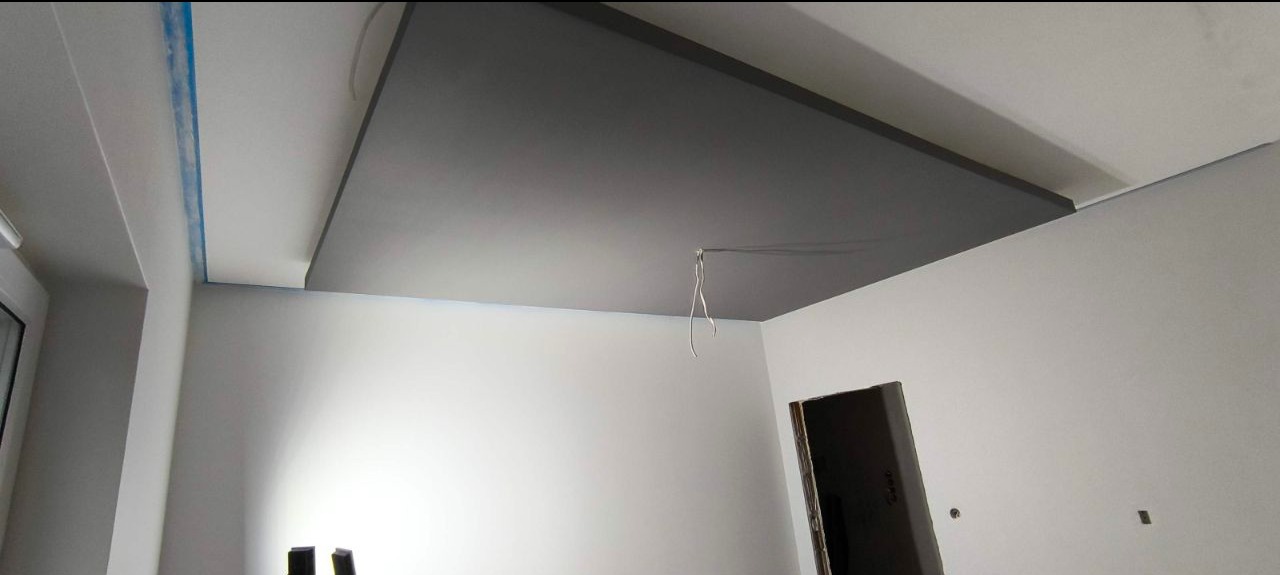 Szara podwieszana płyta karton-gips na suficie w trakcie montażu, widoczne kable i taśma malarska. Ściany pomalowane na biało. Ujęcie z dołu, szeroki kąt.