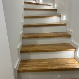 Białe schody z drewnianymi stopniami, oświetlone punktowo, prowadzące do góry. Widok z dołu, ukazujący detale wykończenia i strukturę drewna.