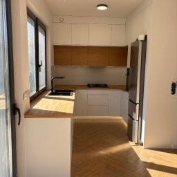 Jasna kuchnia z drewnianymi akcentami: blaty i górne szafki z naturalnego drewna, białe fronty dolnych szafek, czarna armatura, parkiet w jodełkę. Nowoczesny design.