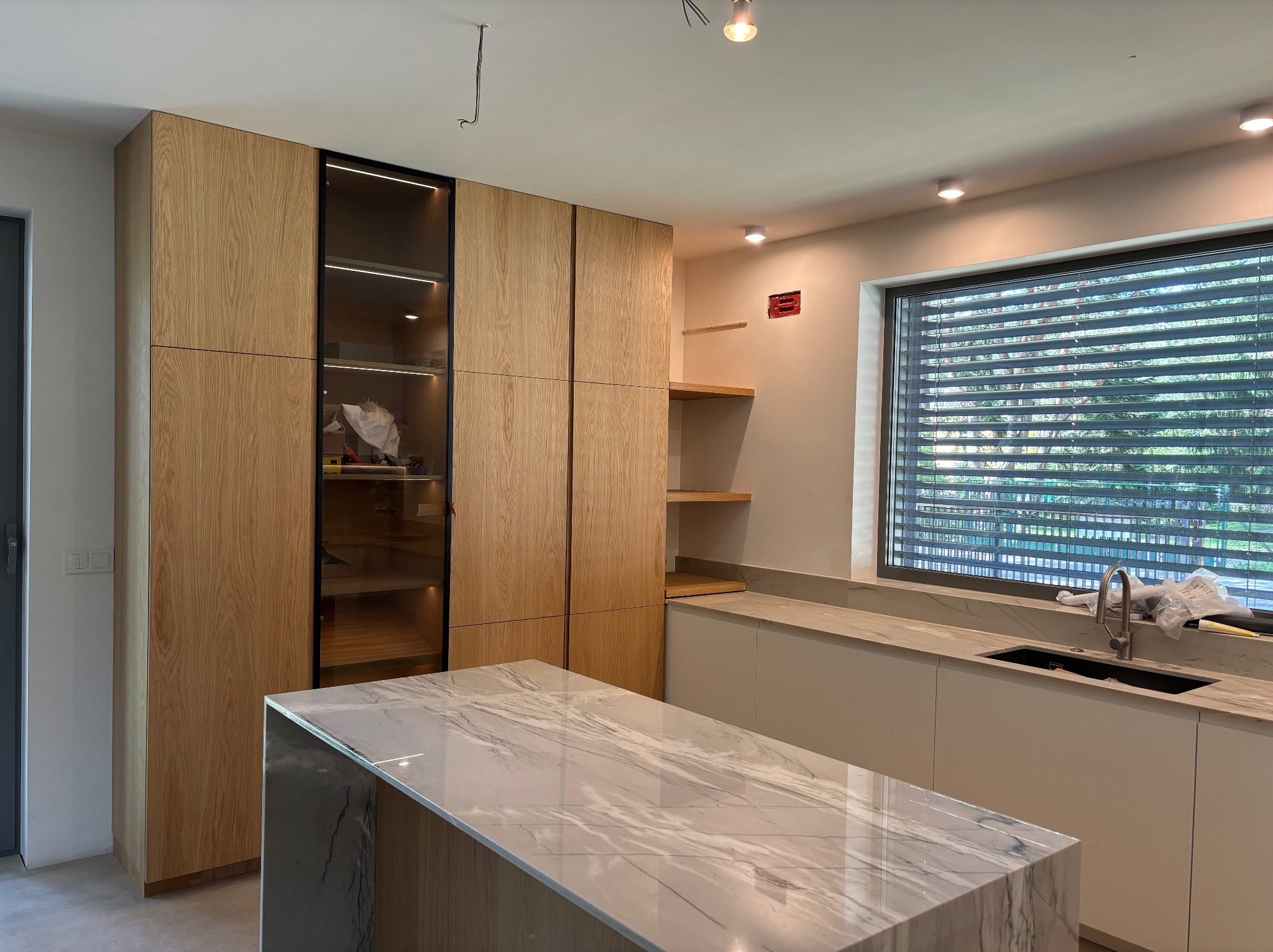 Jasna kuchnia z drewnianymi szafkami i wyspą z marmurowym blatem. Szafka z przeszkleniami i oświetleniem LED. Nowoczesny design, minimalistyczne wykończenie.
