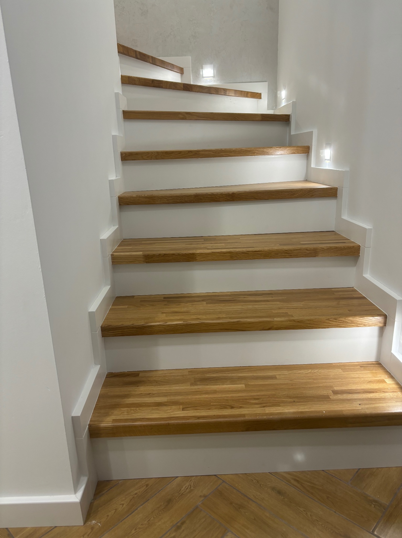 Białe schody z drewnianymi stopniami, oświetlone punktowo, prowadzące do góry. Widok z dołu, ukazujący detale wykończenia i strukturę drewna.