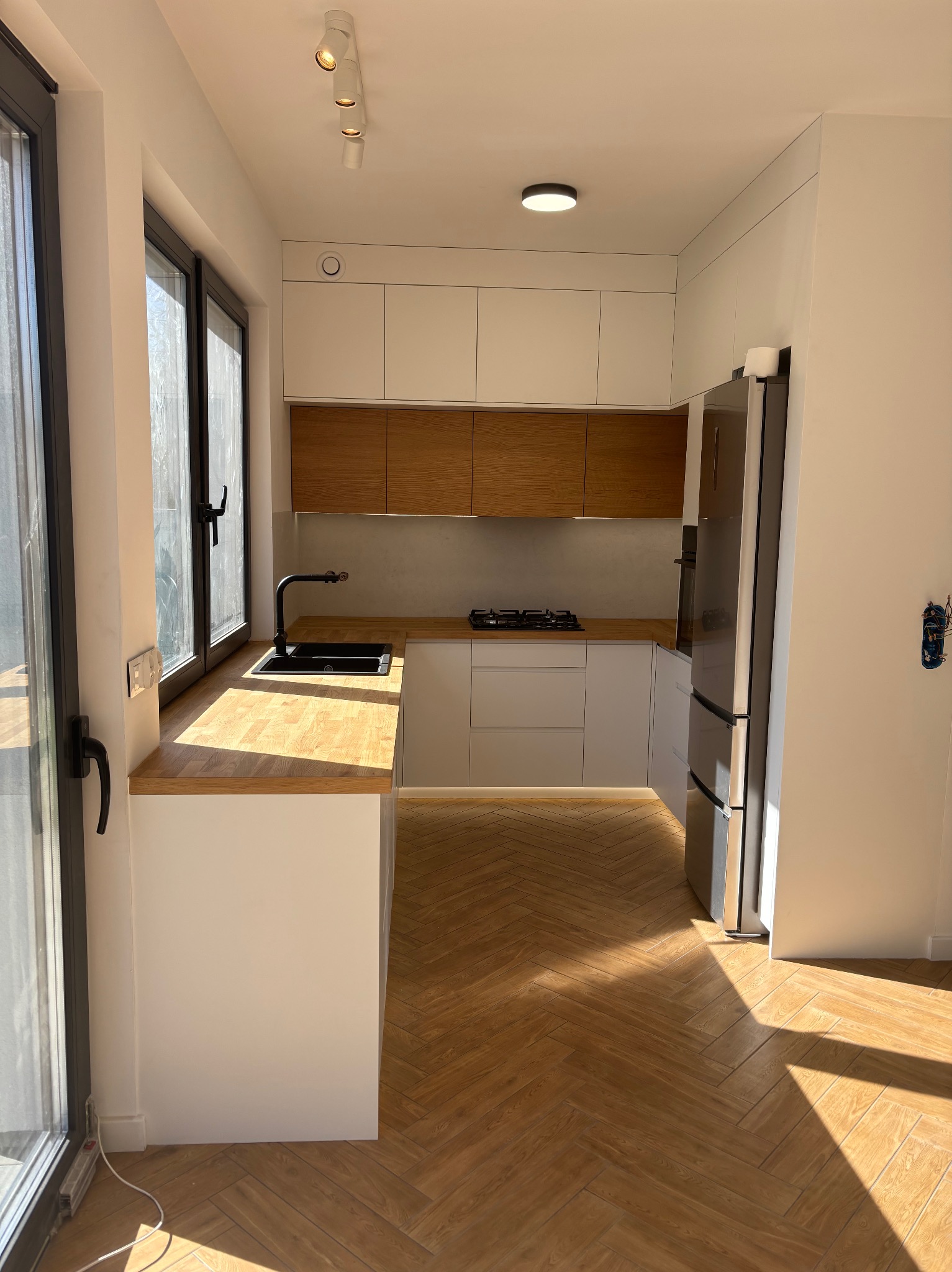 Jasna kuchnia z drewnianymi akcentami: blaty i górne szafki z naturalnego drewna, białe fronty dolnych szafek, czarna armatura, parkiet w jodełkę. Nowoczesny design.