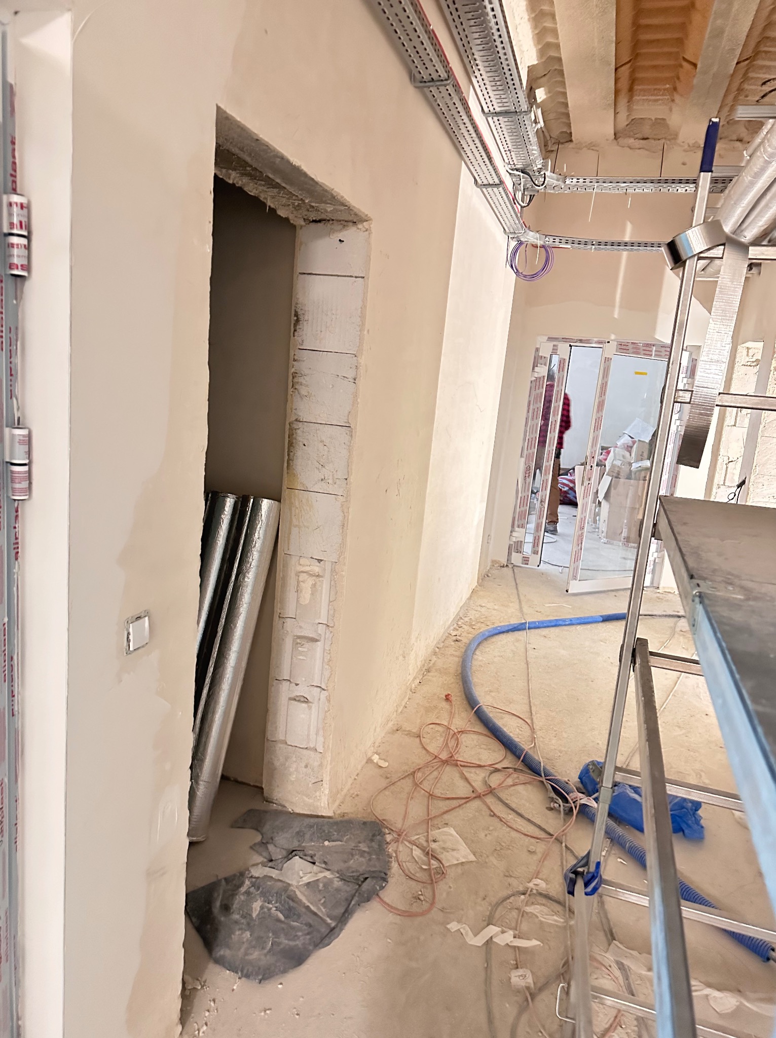 Wnętrze w trakcie remontu: widoczne przewody elektryczne pod sufitem, przygotowane ościeżnice drzwiowe i aluminiowe profile okienne. Na podłodze leżą kable i narzędzia.