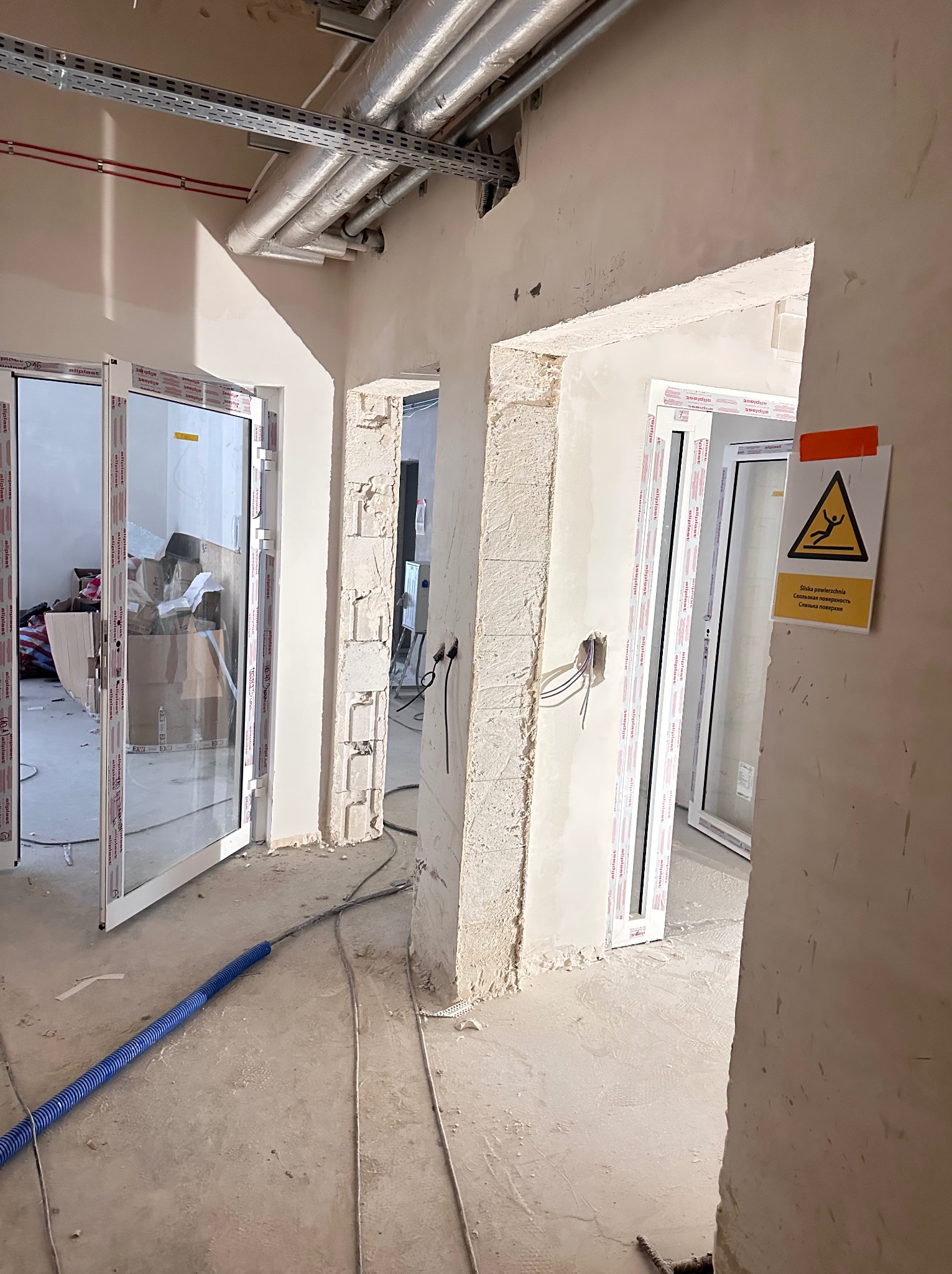 Wnętrze w trakcie remontu: widoczne otwory drzwiowe z zamontowanymi ościeżnicami, instalacje pod sufitem, przewody elektryczne i ostrzeżenie o śliskiej powierzchni.
