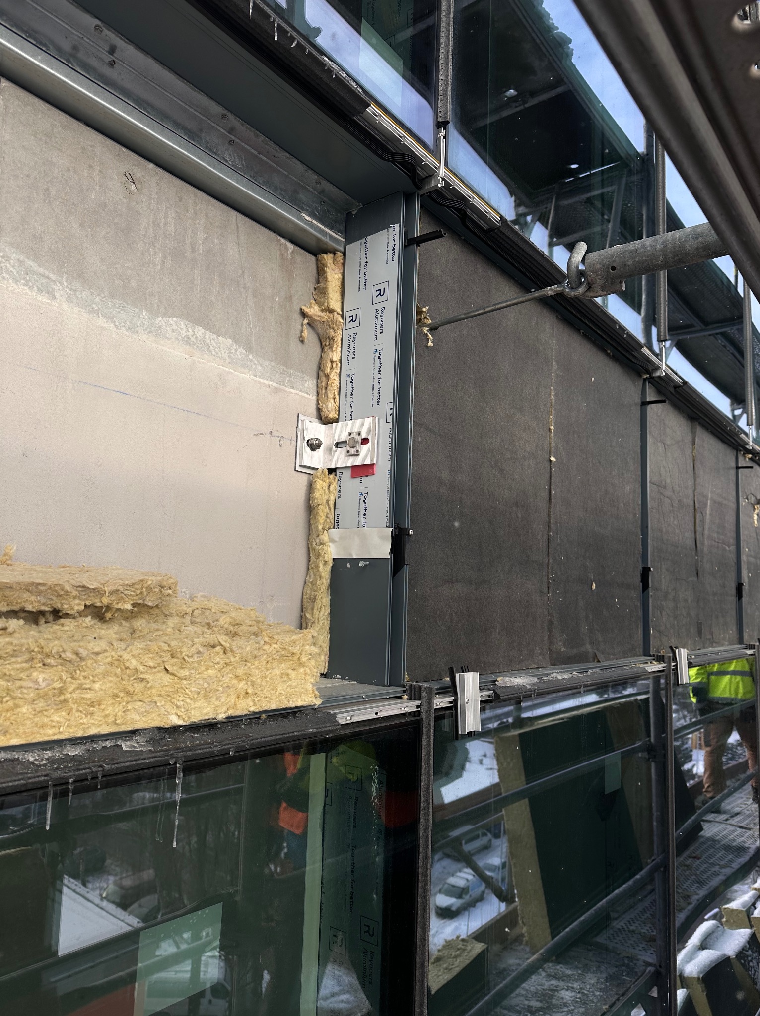 Montaż okien aluminiowych z widoczną izolacją i elementami mocującymi. Widoczne rusztowanie i częściowo zamontowane okna. Prace w toku, detale konstrukcyjne.