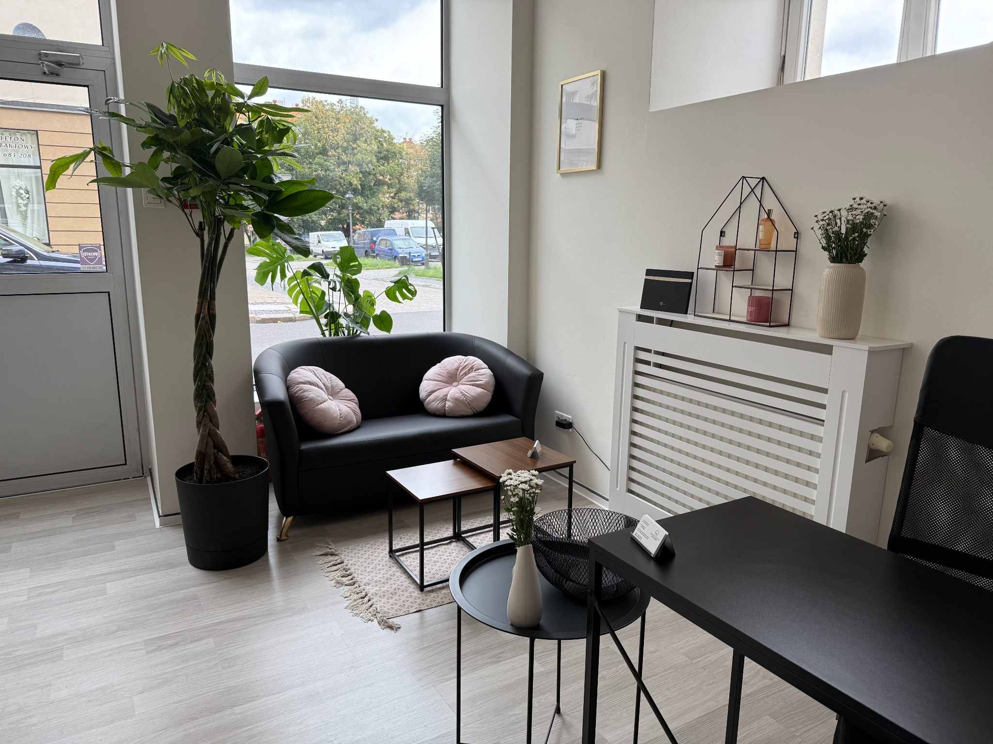 Jasne biuro nieruchomości z czarną sofą, stolikami i roślinami. W tle widok na ulicę przez duże okno. Minimalistyczny wystrój, komfortowa przestrzeń dla klientów.
