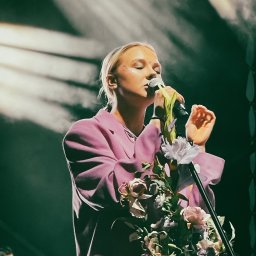 Namika Media Oliwia Nowicka - Piosenkarka w różowym garniturze śpiewa do mikrofonu ozdobionego kwiatami na scenie oświetlonej reflektorami. W tle widoczny muzyk z gitarą.