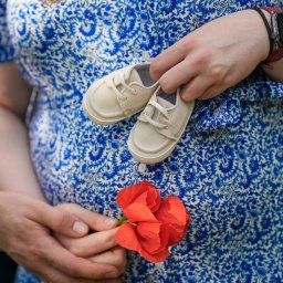 Namika Media Oliwia Nowicka - Brzuch kobiety w kwiecistej sukience, trzymającej małe buciki i czerwony mak. Ujęcie z sesji ciążowej, skupiające się na symbolice oczekiwania na dziecko.