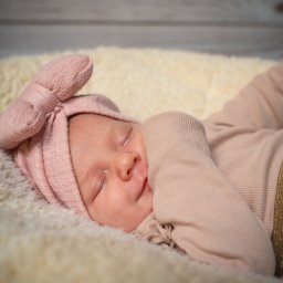 Namika Media Oliwia Nowicka - Śpiąca noworodka w różowym ubranku i opasce z kokardą, leżąca na puszystym, kremowym kocyku na tle drewnianej podłogi. Delikatne światło podkreśla rysy twarzy.