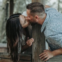 Namika Media Oliwia Nowicka - Zakochana para całuje się, opierając się o drewnianą konstrukcję na tle zieleni. Kadr z sesji narzeczeńskiej, delikatne światło, naturalna sceneria.
