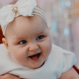 Namika Media Oliwia Nowicka - Uśmiechnięta niemowlęca dziewczynka z niebieskimi oczami i białą koronkową opaską na głowie, trzymana na rękach, w tle rozmyte dekoracje.