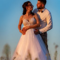 Namika Media Oliwia Nowicka - Para młoda w objęciach na tle błękitnego nieba i łąki. Panna młoda w okularach i sukni z koronkową górą, pan młody w muszce. Romantyczna sesja w plenerze.