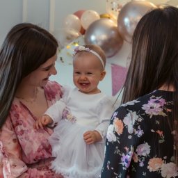 Namika Media Oliwia Nowicka - Uśmiechnięta dziewczynka w białej sukience na tle balonów, trzymana przez kobietę w Bogdanowie. Radosna atmosfera, jasne oświetlenie, dekoracje urodzinowe.